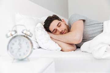 Photo of handsome man having stubble and wearing casual clothes, sleeping at home in bed with clock...