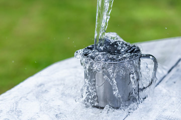 A steel mug with water and sprays on a white wooden table on nature background, life concept Water is pouring  out of the bucket into the mug on a white wooden table on nature background