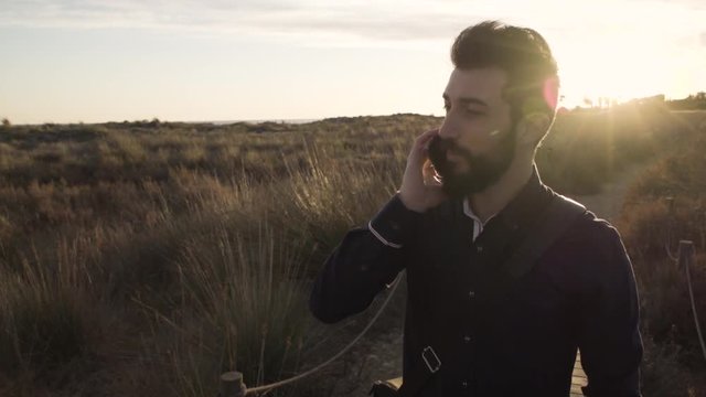 Content adult man with beard in stylish outfit speaking on phone against countryside in sunlight.