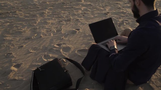 Adultbusiness man sitting ona beach and using laptop working remotely.
