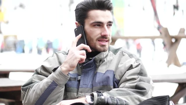 Handsome young man in skiing suit relaxing, using cell phone to make a call, sitting at table outside ski resort