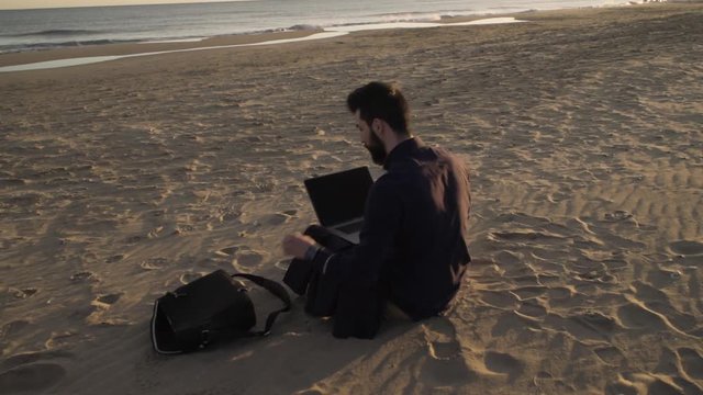 Concentrated man relaxing on a beach browsing laptop in sunlight.