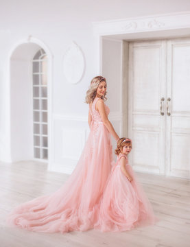 Mom and daughter in luxurious, pink dresses with a long train. Family clothes, identical dresses. The background is beautiful, expensive, white, classic interior. Artistic, family photography