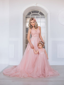 Mom and daughter in luxurious, pink dresses with a long train. Family clothes, identical dresses. The background is beautiful, expensive, white, classic interior. Artistic, family photography