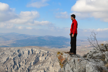 Young man on a cliff edge on the top of mountain with gorgeous view. Hiker on mountain peak on beautiful sunny day