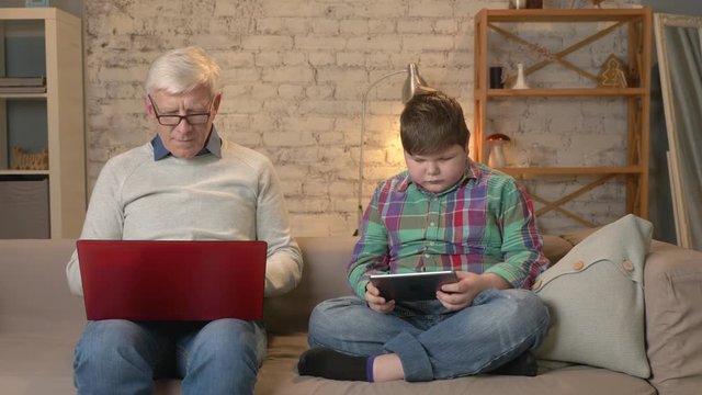 The difference of generations. Elderly man with glasses sitting on sofa working on laptop, young fat boy playing on tablet. Home comfort, family idyll, cosiness concept 60 fps