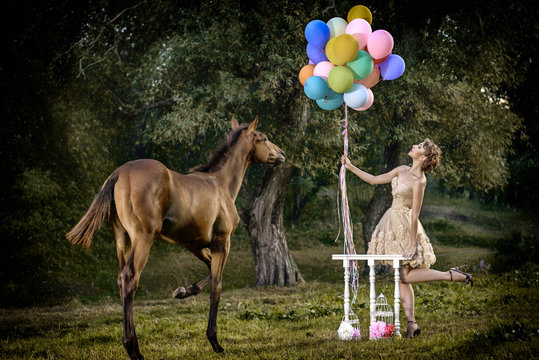 Art photo outdoor in fairytale forest. Beautiful slim model woman with short brown curly hair in wonderful dress cute posing smiling with colorful balloons on the meadow with amazing slender horse