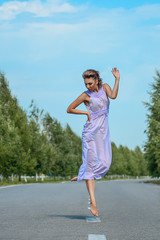 Beautiful slim model woman with short brown curly hair in a purple transparent silk dress gently posing in dancing jump on the road with shiny sun, blue sky and green trees. Fashion photo shooting