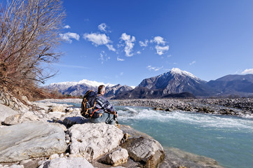 Hiker sitting on river shore, looking the mountains landscape.