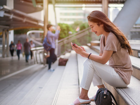 Young Asian Woman Sitting And Smiling, Using Smartphone Searching For Social Media In The City.