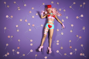 lebende Barbiepuppe in knappen blauen Lackhotpants und high heels liegt träumend inmitten von vielen Lollypops auf einem knallig Violetten  Hintergrund.
