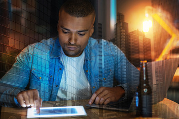 Serious man. Calm attentive young man looking concentrated while sitting at the table with a modern laptop by his side and touching the screen of a tablet in front of him