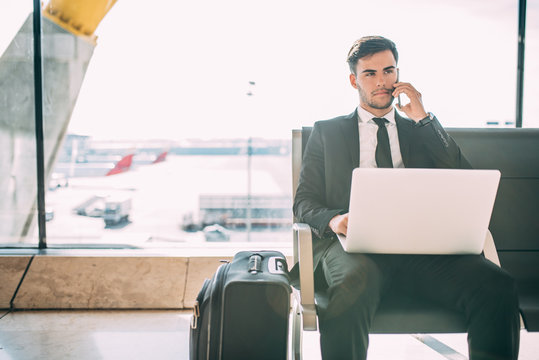 Young business man sitting on the computer and mobile phone with the suitcase at the airport waiting for the flight
