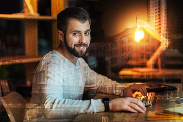 Optimistic man. Calm handsome smiling man sitting comfortably at the table with his hands on the keyboard and feeling satisfied after a long productive working day