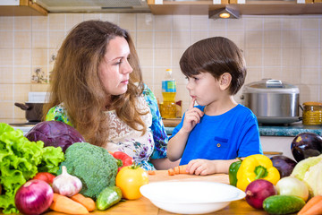 The young cook mother standing with her little son in the kitchen and salting vegetables