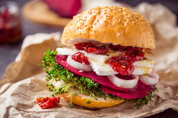 Vegitarian burger with beet slices, microgreen sprouts, tofu cheese, dried tomatoes and onion on kraft package paper on dark stone background with inhrediants. Soft selective focus. Copy space.