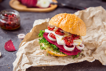Vegitarian burger with beet slices, microgreen sprouts, tofu cheese, dried tomatoes and onion on kraft package paper on dark stone background with inhrediants. Soft selective focus. Copy space.