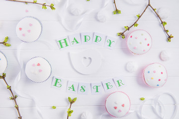 Top view holiday composition of Happy Easter lettering, branches with young shoots of greenery, decorated cupcakes, merengue sweets, bird figure on wooden background. Art concept. Selective focus.
