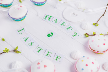 Obraz na płótnie Canvas Holiday composition of Happy Easter lettering, branches with young shoots of greenery, decorated cupcakes, merengue sweets, bird figure on wooden background. Art concept. Selective focus.