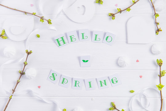 Top view composition of Hello spring lettering, branches with young shoots of greenery, merengue sweets and candy hearts, handcraft bird figure on wooden background. Art concept. Selective focus.