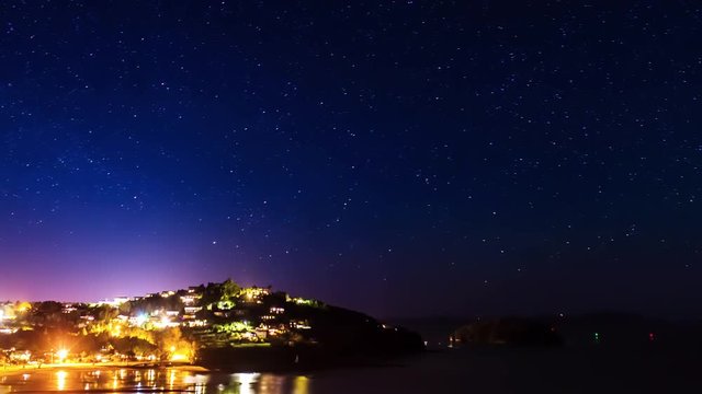 Time lapse of a starry sky above a small city illuminated in front of the sea and a beach