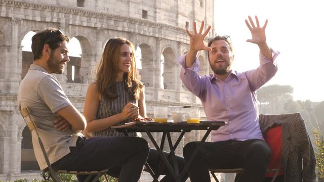 Three young friends having fun laughing telling stories and jokes with exagerated gestures sitting at bar restaurant table in front of colosseum in rome at sunset