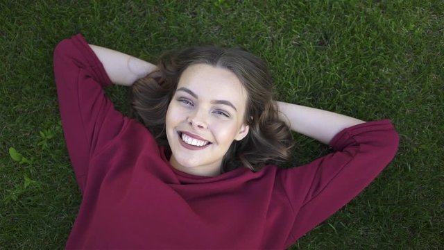 Top view of a cheerful young woman wearing a red sweater and lying on the grass with her hands behind her head. Handheld real time medium shot