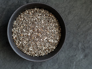 Chia seeds in a black metal bowl on a black textured board. Top view with copy space.