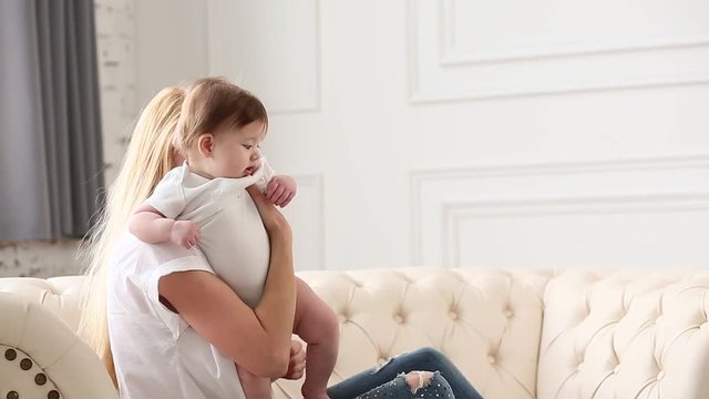 Pretty young woman holding baby girl in her arms at domestic room at the morning. Happy mother hugging and kissing her little newborn daugther at home