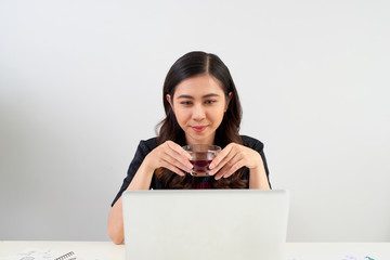 Young woman taking a tea break as she sits at a table working on a laptop computer,