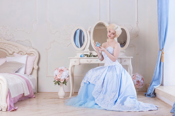 A beautiful, young blonde woman sits by the dressing table and holds a blue mask dressed in a ball...