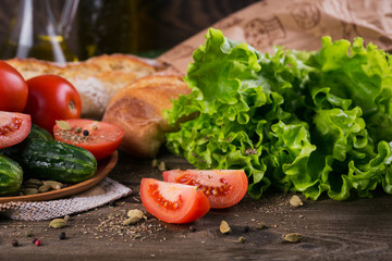 Fresh ingredients for cooking salad: tomato, cucumber, lettuce, olive oil and spices over rustic wooden table background. Served with fresh baguette. Healthy eating concept