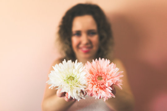 Beautiful curly young woman holding two flowers, pink and white on color background at home. Hello Spring concept. Focus on the flowers.