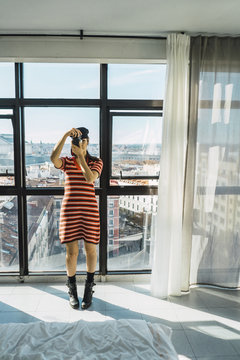 Full length of young woman photographing with camera while standing against windows at home