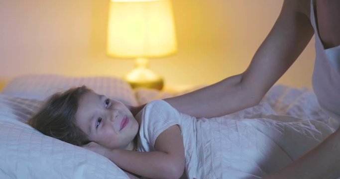 Goodnight mother to her little daughter who sleeps at night in her bedroom and sees the moonlight coming through the window and lighting her sweet face. Concept of: peaceful dreams, family, relaxation