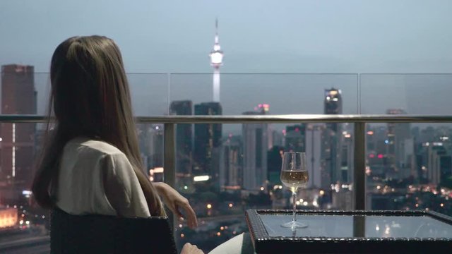 Happy woman drinking a glass of white wine while sitting in rooft bar in the city or on the terrace with cityscape at sunset. Vacation,happiness,freedom concept.