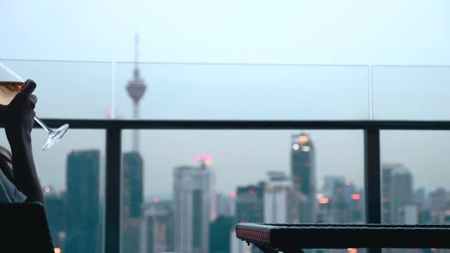 Happy woman drinking a glass of white wine while sitting in rooft bar in the city or on the terrace with cityscape at sunset. Vacation,happiness,freedom concept.