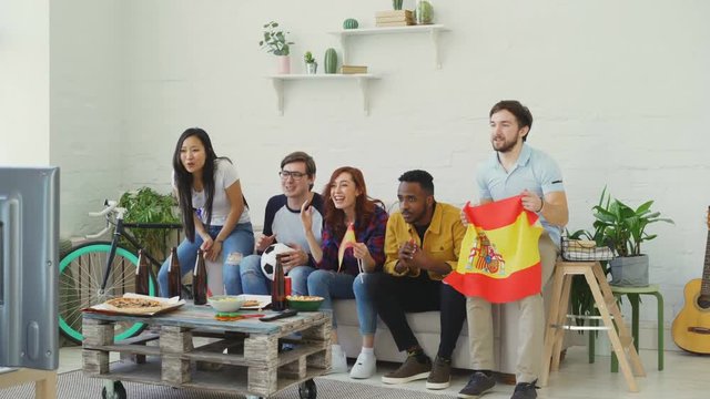 Multi-ethnic group of friends sports fans with Spanish flags watching football championship on TV together at home and cheering up favourite team