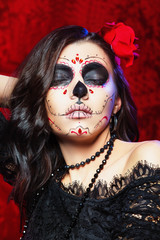 Vertical portrait of traditional mexiacan halloween face art. Brunette lady with rose in hair at red background.