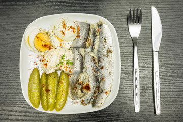 White plate with marinated herring in oil, boiled hard-boiled egg, cucumber and mayonnaise. Seasoned with black pepper and red paprika. View from above.