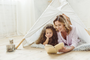 happy mother and daughter reading book together in kid wigwam