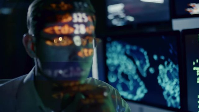 Concentrated young IT professional in eyeglasses looking at monitor in dark office at night; computer data projected on his face