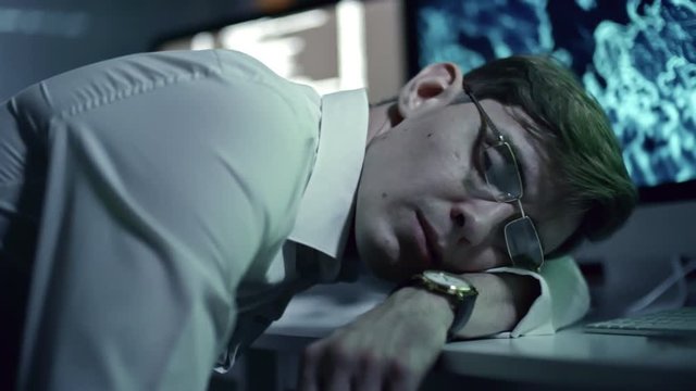 Tracking shot of tired office worker in eyeglasses sleeping on desk in front of computer in cyber security center at night
