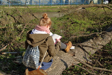 nastolatka z blond włosami spiętymi w kok siedzi na ściętym drzewie w parku, czyta otwartą grubą książkę, ubrana w zielona kurtkę, spodnie, różowy szalik, ma młodzieżowy plecak, w ścięte drzewa 