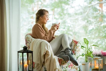 Foto op Plexiglas Young woman sitting home in a chair by the window with cup of hot coffee wearing knitted warm sweater. Cozy room decorated with lanterns and candles. Scenic view of pine trees in snow in window © Suzi Media 