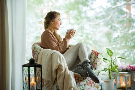 Young Woman Sitting Home In A Chair By The Window With Cup Of Hot Coffee Wearing Knitted Warm Sweater. Cozy Room Decorated With Lanterns And Candles. Scenic View Of Pine Trees In Snow In Window