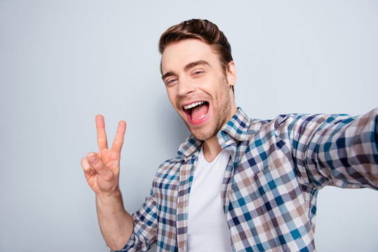 Self portrait of bearded, cool, mad, brunet guy in casual outfit, checkered shirt showing v-sign to the camera, laughing, shouting, screaming over grey background