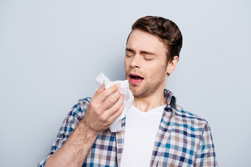 Portrait of attractive, handsome, stylish guy in checkered shirt, sneezing with open mouth and close eyes, holding tissues for his runny nose, having a cold, standing over grey background