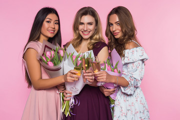 Three beautiful girls in colorful  dresses are holding tulip bouquets. Ladies are drinking champagne. Pink background.