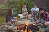 Company of young people holding sticks with marshmallows above campfire in the evening